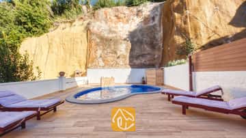 Piscina privada en Villa Blanca con azulejos de mosaico azul, ubicada en un patio adoquinado bordeado por una pared de roca alta y tumbonas de madera con cojines morados.