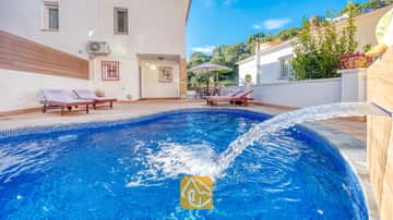 Piscina curva de azulejos azules con fuente en Villa Blanca, que cuenta con tumbonas de madera en el patio pavimentado junto a una pared exterior blanca.