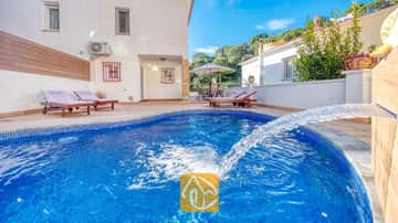 Piscina curva de azulejos azules con fuente en Villa Blanca, que cuenta con tumbonas de madera en el patio pavimentado junto a una pared exterior blanca.