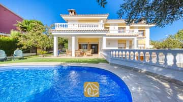 Piscina curva de azulejos azules en primer plano, con la Villa Baileys de exterior blanco, paredes amarillas y balcones alicatados bajo un cielo despejado.