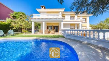 Piscina curva de azulejos azules en primer plano, con la Villa Baileys de exterior blanco, paredes amarillas y balcones alicatados bajo un cielo despejado.