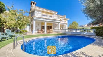 Piscina privada de aguas azuladas con pasamanos metálicos frente al exterior de la Villa Baileys de dos plantas, que cuentaกับ pilares blancos, balcones y un tejado de tejas bajo un cielo despejado.