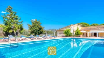 Piscina al aire libre en Villa Miro con tumbonas azules y blancas sobre una terraza de terracota. Árboles verdes y un edificio amarillo se alzan bajo un cielo azul despejado.