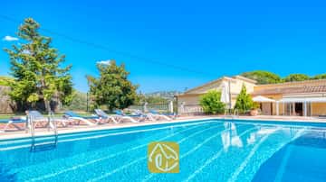 Piscina al aire libre en Villa Miro con tumbonas azules y blancas sobre una terraza de terracota. Árboles verdes y un edificio amarillo se alzan bajo un cielo azul despejado.