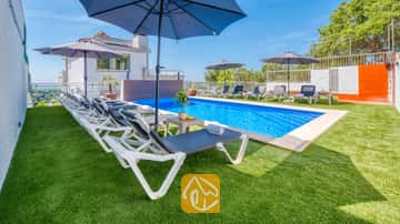 Piscina privada en Villa Sandy Beach con hileras de tumbonas blancas, sombrillas azules y césped artificial verde bajo un cielo despejado.