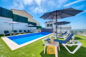 Piscina privada en Villa Sandy Beach rodeada de césped artificial, tumbonas grises con estructura blanca y grandes sombrillas bajo un cielo azul despejado.