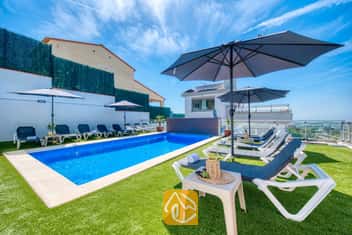 Piscina privada en Villa Sandy Beach rodeada de césped artificial, tumbonas grises con estructura blanca y grandes sombrillas bajo un cielo azul despejado.