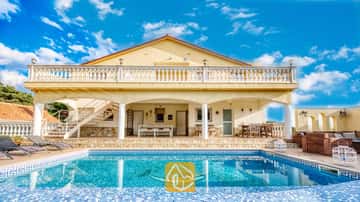 Vista exterior de Villa Madonna, que cuenta con una piscina con un mosaico de delfines, una veranda sombreada, un patio revestido de piedra y paredes exteriores de color amarillo bajo un cielo azul.