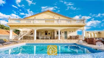 Vista exterior de Villa Madonna, que cuenta con una piscina con un mosaico de delfines, una veranda sombreada, un patio revestido de piedra y paredes exteriores de color amarillo bajo un cielo azul.