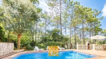 Piscina privada en la Villa Esmee con tumbonas blancas en el patio enlosado, rodeada de altos pinos y una barbacoa de piedra.