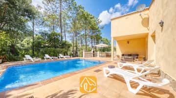 Piscina exterior privada en Villa Esmee con tumbonas blancas en un patio de losetas, rodeada de pinos bajo un cielo azul.