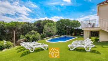 Piscina al aire libre rodeada de césped verde y tumbonas blancas en Villa La Luna, España, con un telón de fondo de árboles y cielo azul.