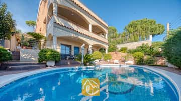 Piscina curva con suelo de azulejos de mosaico situada frente a la fachada de varias plantas de Villa Grace, que cuenta con arcos de piedra y balcones bajo un cielo azul despejado.