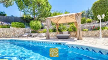 Piscina privada en Villa Lorena con bordes de azulejos azules, una pérgola de lona beis sobre la zona de estar exterior, muros de contención de piedra y vegetación alrededor.
