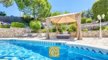 Piscina privada en Villa Lorena con bordes de azulejos azules, una pérgola de lona beis sobre la zona de estar exterior, muros de contención de piedra y vegetación alrededor.