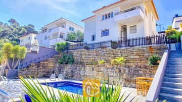 Vista exterior de Villa Fransisca que muestra una piscina rectangular junto a muros de contención de piedra en terrazas, bajo un edificio blanco de varias plantas y un cielo azul.