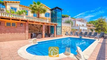 Piscina privada y terraza en Villa Dolce Vita, que cuenta con un hueco de ascensor de cristal, paredes de ladrillo y numerosas tumbonas bajo un cielo azul.