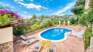 Piscina privada con tumbonas y terraza de baldosas en Villa Cleo, España, rodeada de muros de piedra y arbustos en flor.