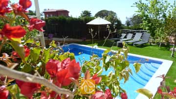 Piscina privada en Villa Noa enmarcada por buganvillas rojas, con tumbonas y una sombrilla blanca en el césped al fondo.