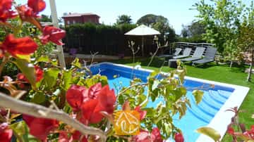 Piscina privada en Villa Noa enmarcada por buganvillas rojas, con tumbonas y una sombrilla blanca en el césped al fondo.