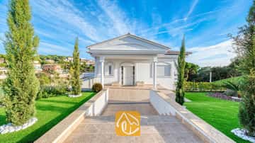 Fachada exterior de color blanco de Villa Madison, que cuenta con una entrada de camino adoquinado, columnas y céspedes verdes a los lados con cipreses bajo un cielo azul.