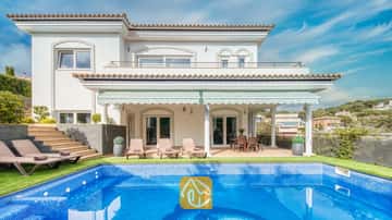 Vista exterior de Villa Madison que muestra una piscina revestida de azulejos azules, tumbonas y una terraza cubierta con columnas blancas bajo un cielo azul despejado.