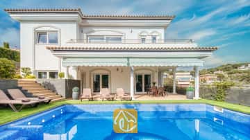 Vista exterior de Villa Madison que muestra una piscina revestida de azulejos azules, tumbonas y una terraza cubierta con columnas blancas bajo un cielo azul despejado.