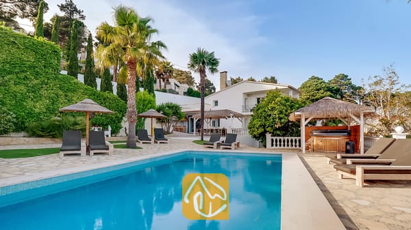 Zona de piscina al aire libre en Villa Tropicana con tumbonas, palmeras, sombrillas de paja y un cenador de madera que alberga una bañera de hidromasaje.
