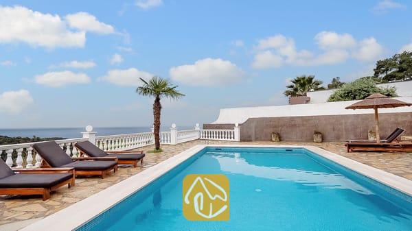 Piscina privada en Villa Tropicana rodeada de suelo de piedra, tumbonas de madera con cojines oscuros y una balaustrada blanca con vistas al mar bajo un cielo azul.