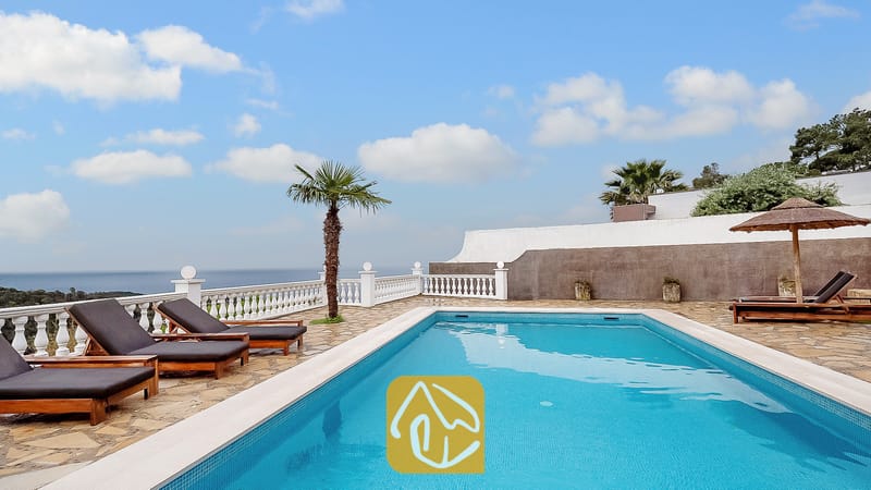 Piscina privada en Villa Tropicana rodeada de suelo de piedra, tumbonas de madera con cojines oscuros y una balaustrada blanca con vistas al mar bajo un cielo azul.