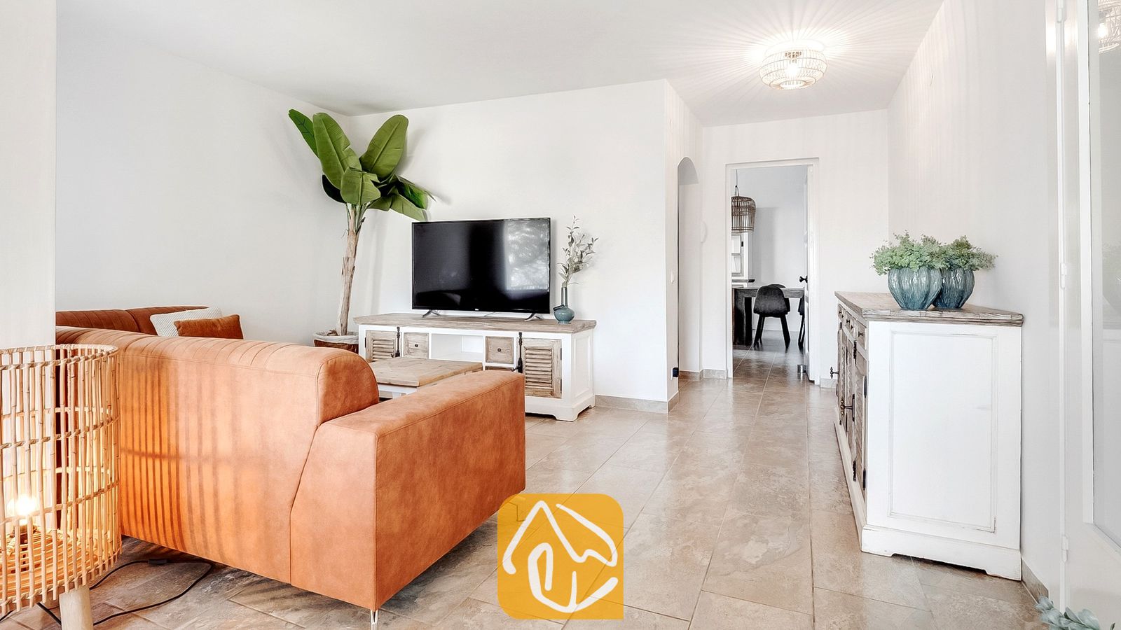 Salón de Villa Tropicana con un sofá de piel marrón, televisión de pantalla plana sobre un mueble multimedia de estilo rústico en color blanco y suelos de baldosa.