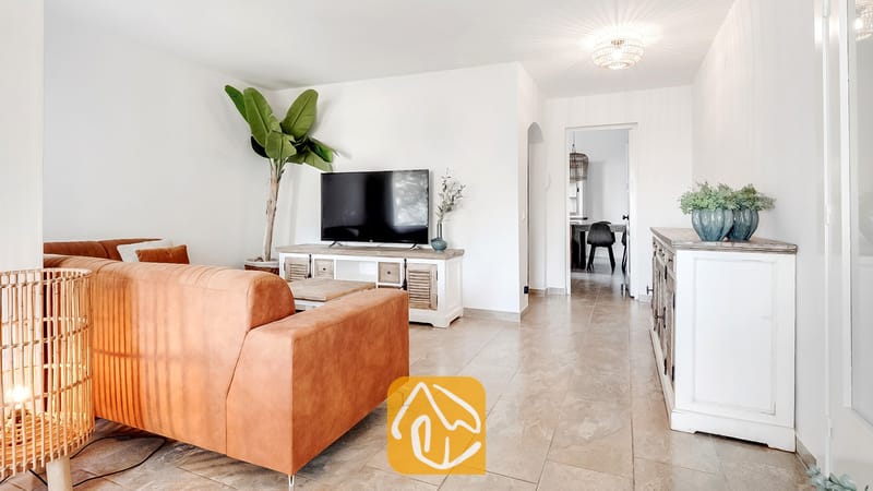 Salón de Villa Tropicana con un sofá de piel marrón, televisión de pantalla plana sobre un mueble multimedia de estilo rústico en color blanco y suelos de baldosa.