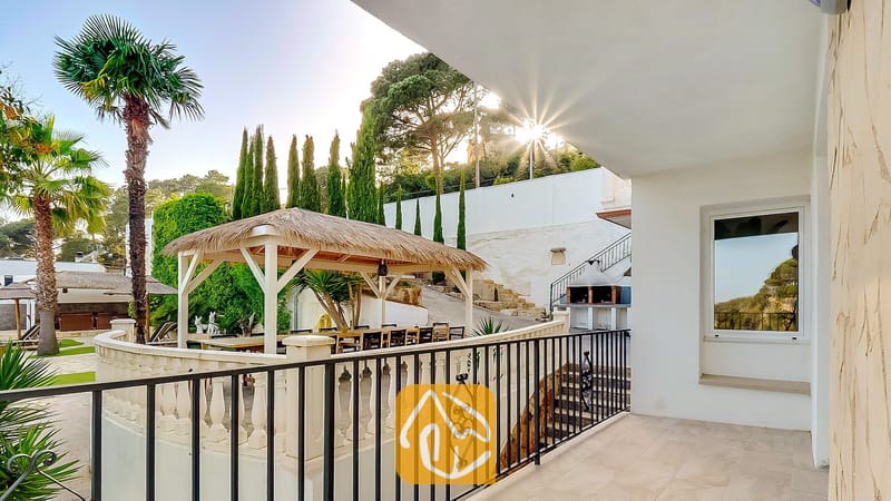 Terraza cubierta en Villa Tropicana con vistas a una palapa, zona de comedor al aire libre, palmeras y una barbacoa de ladrillo junto a la ladera.