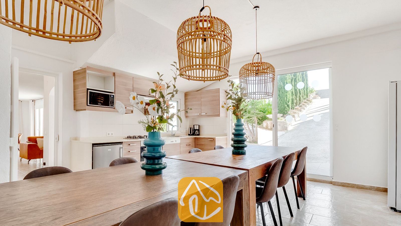 Mesa de comedor de madera con sillas oscuras y jarrones turquesas, situada bajo lámparas colgantes de mimbre tejido en el interior de Villa Tropicana, con una cocina visible al fondo.