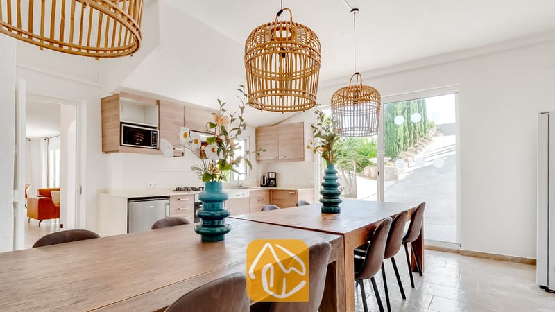 Mesa de comedor de madera con sillas oscuras y jarrones turquesas, situada bajo lámparas colgantes de mimbre tejido en el interior de Villa Tropicana, con una cocina visible al fondo.