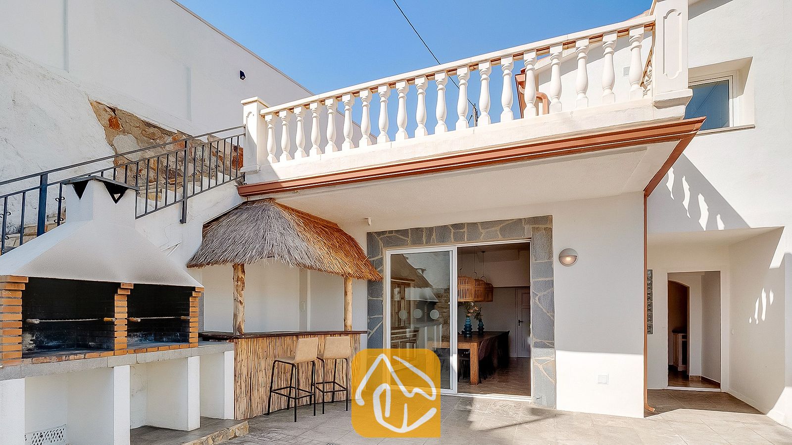 Terraza exterior en Villa Tropicana que cuenta con una barbacoa de obra de ladrillo, un bar tiki con techo de paja y dos taburetes, y una barandilla de balcón blanca.