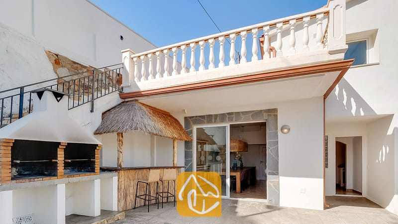 Terraza exterior en Villa Tropicana que cuenta con una barbacoa de obra de ladrillo, un bar tiki con techo de paja y dos taburetes, y una barandilla de balcón blanca.