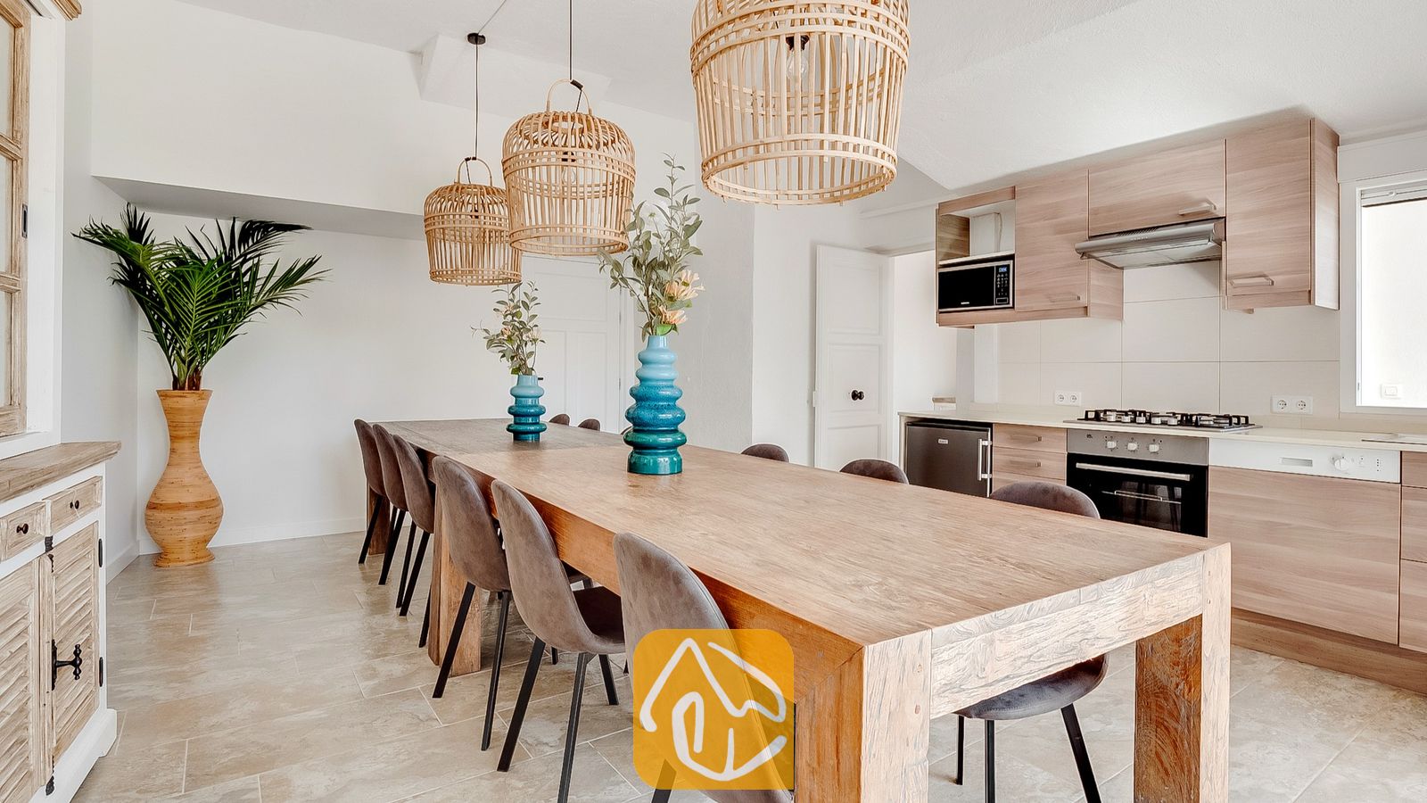 Villa Tropicana cuenta con una cocina abierta y un comedor equipado con una larga mesa de madera, sillas tapizadas en gris, lámparas colgantes de mimbre y gabinetes de madera clara.