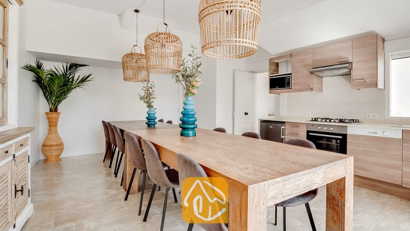 Villa Tropicana cuenta con una cocina abierta y un comedor equipado con una larga mesa de madera, sillas tapizadas en gris, lámparas colgantes de mimbre y gabinetes de madera clara.