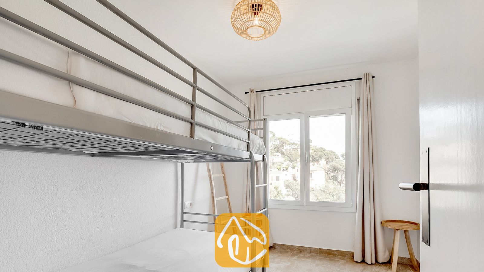 Dormitorio con literas de metal gris, paredes blancas y una ventana con cortinas ligeras que ofrece vistas a los árboles.