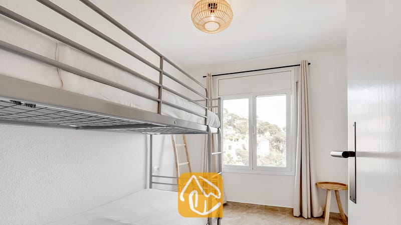 Dormitorio con literas de metal gris, paredes blancas y una ventana con cortinas ligeras que ofrece vistas a los árboles.