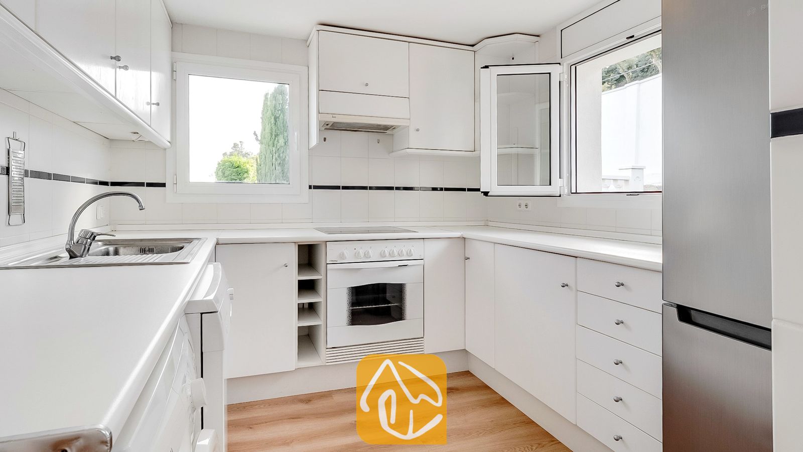 Cocina blanca y luminosa equipada con gabinetes blancos, horno empotrado con placa de cocción, refrigerador de acero inoxidable y dos ventanas con vistas a los árboles.