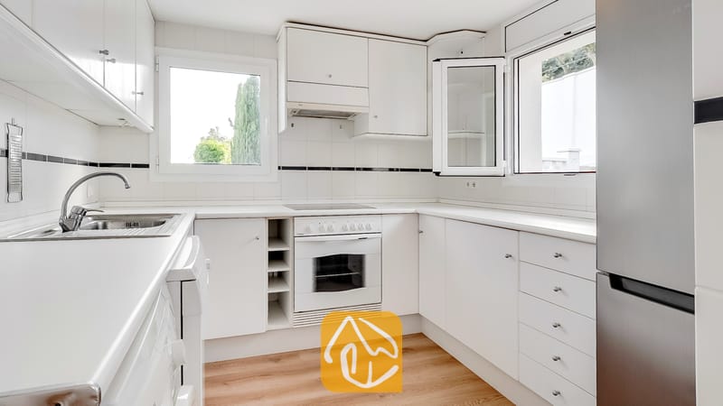 Cocina blanca y luminosa equipada con gabinetes blancos, horno empotrado con placa de cocción, refrigerador de acero inoxidable y dos ventanas con vistas a los árboles.