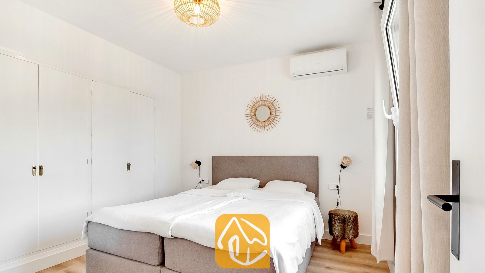 Dormitorio con cama doble tapizada en gris, ropa de cama blanca y un espejo redondo de mimbre con diseño de sol en la pared blanca.