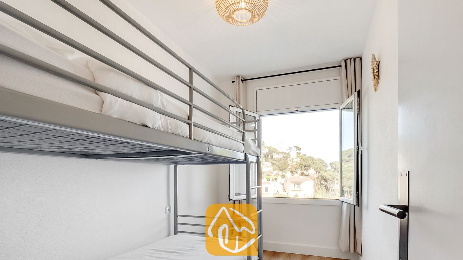 Dormitorio con literas de metal vestidas con ropa de cama blanca, una lámpara de techo tejida y una ventana abierta con vistas al verde exterior.