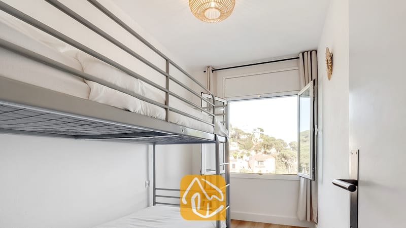Dormitorio con literas de metal vestidas con ropa de cama blanca, una lámpara de techo tejida y una ventana abierta con vistas al verde exterior.