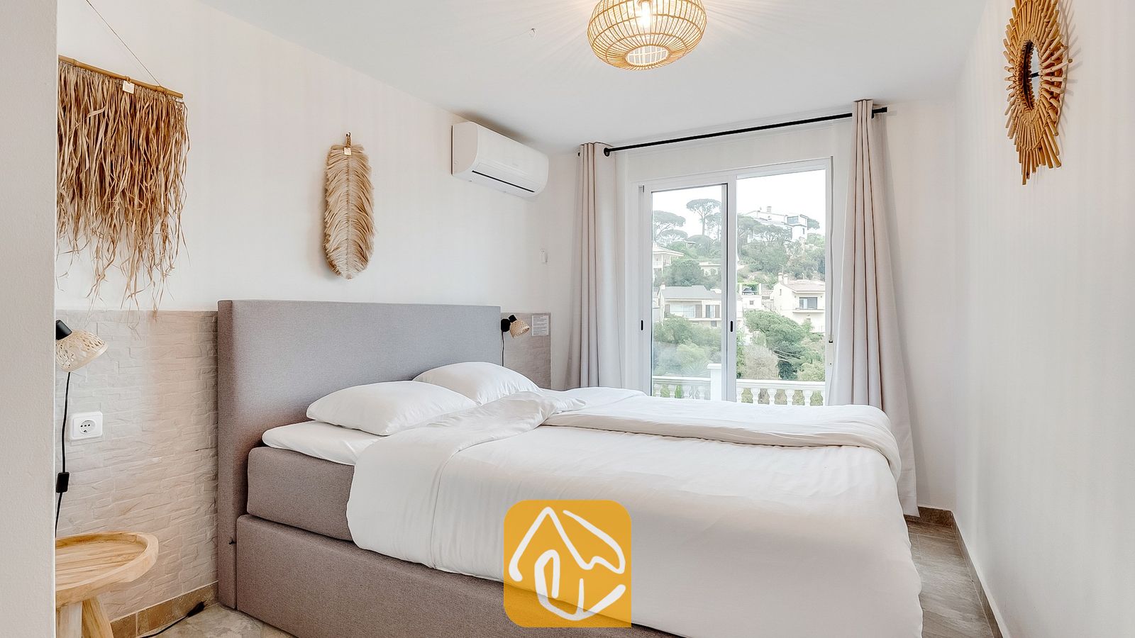 Dormitorio con una cama tapizada en gris, sábanas blancas y puertas corredizas de vidrio que se abren a una vista verde de la colina.