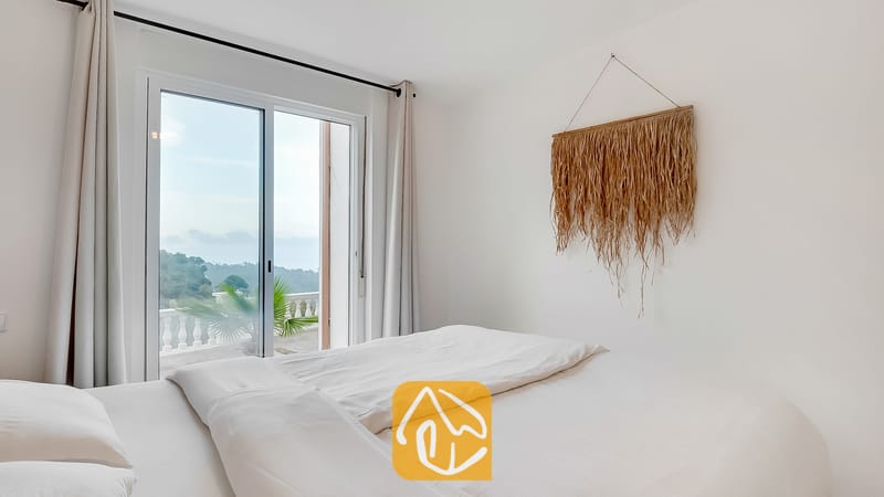 Dormitorio con ropa de cama blanca, un tapiz de pared de paja tejida y puertas corredizas de vidrio que se abren a una terraza con vistas verdes.