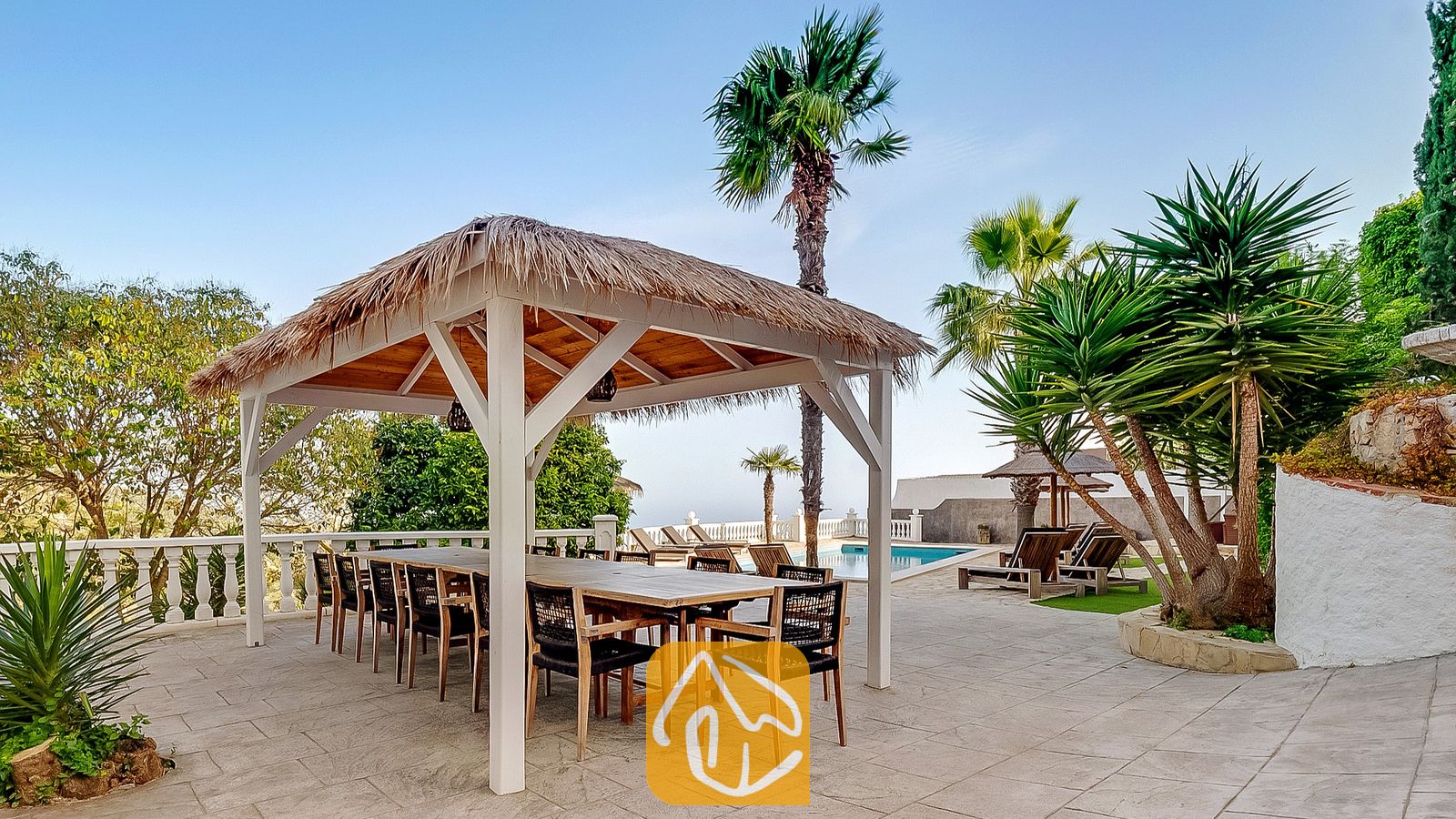Pérgola de comedor exterior cubierta con techo de paja y una gran mesa de madera en Villa Tropicana, situada en una terraza de azulejos junto a palmeras y una piscina.