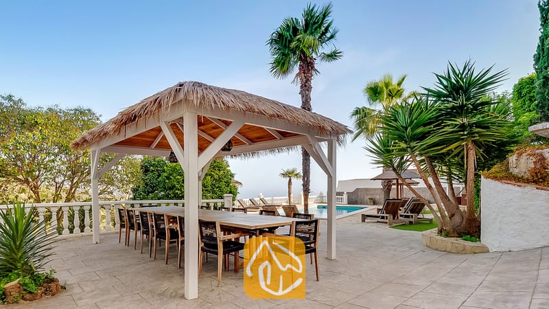 Pérgola de comedor exterior cubierta con techo de paja y una gran mesa de madera en Villa Tropicana, situada en una terraza de azulejos junto a palmeras y una piscina.